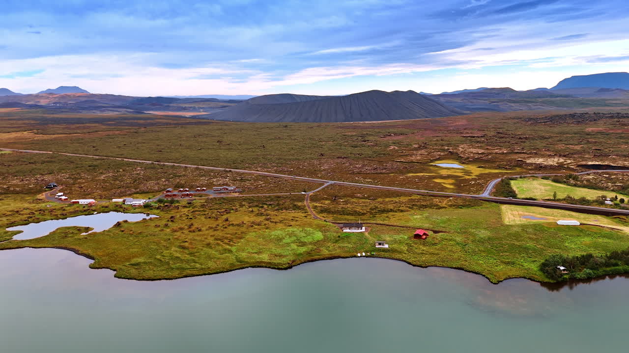 Approaching plain waterscape with some houses and highways on. Huge volcano crater at backdrop. Aerial perspective on Iceland scenery.