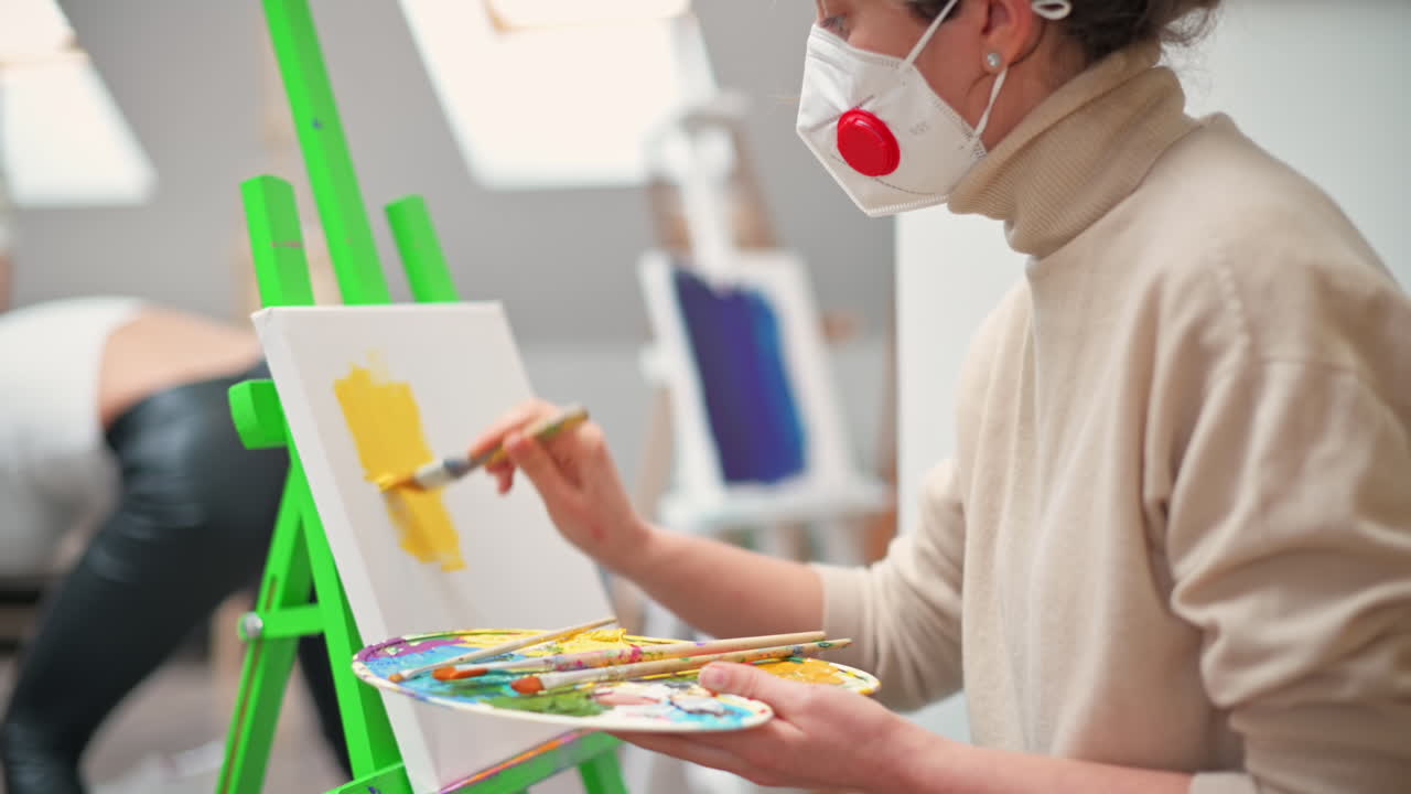 A woman in medical mask painting a picture using a palette and brush in a studio