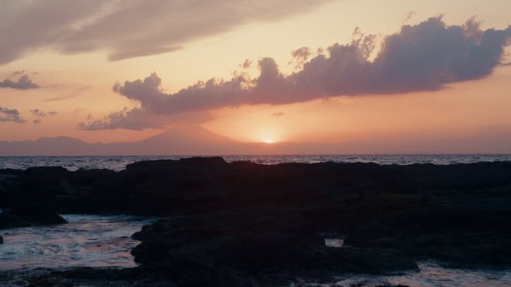 Fiery Sunset on Japan’s Shore – Stunning waves lap dark rocks as Mount Fuji rests in the distant glow.
