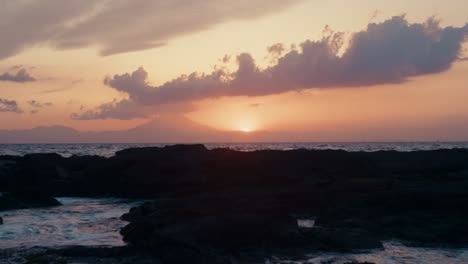fiery sunset on japan’s shore – stunning waves lap dark rocks as mount fuji rests in the distant glow.