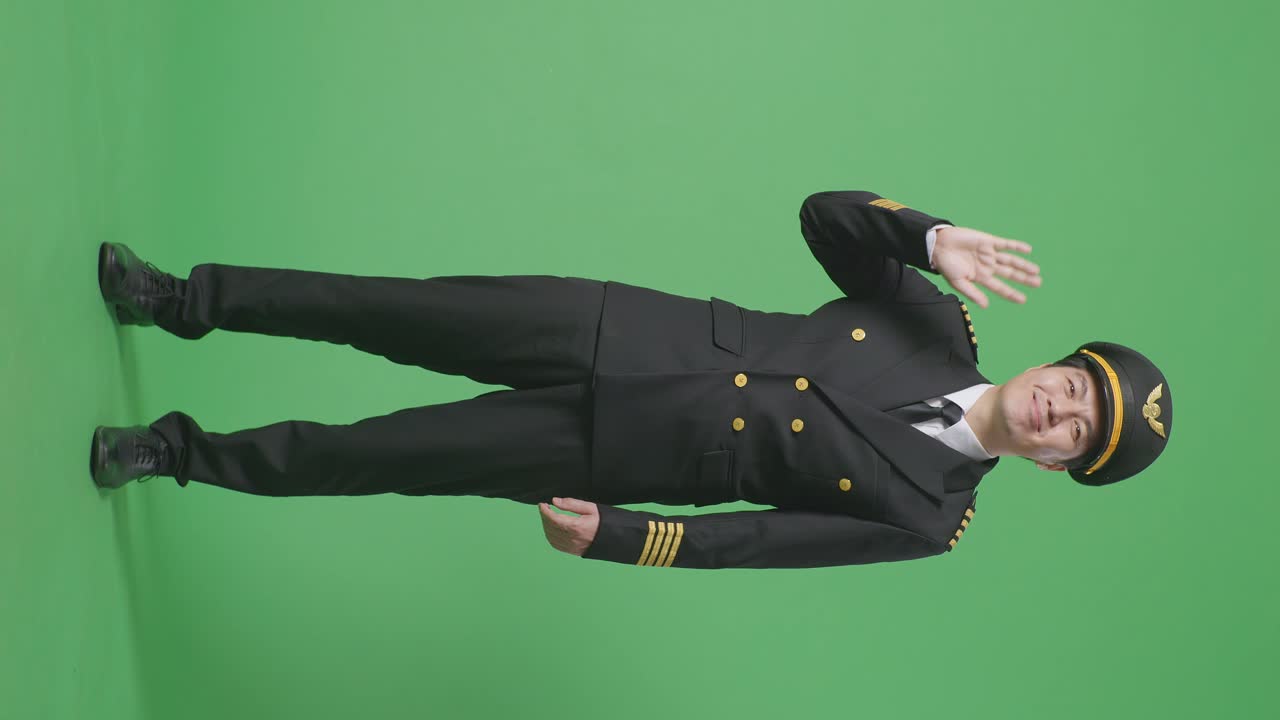 아시아 남성 조종사가 미소 짓고, 손을 흔들며, 녹색 스크린 배경 스튜디오에 서서 작별 인사를 하고 있습니다.