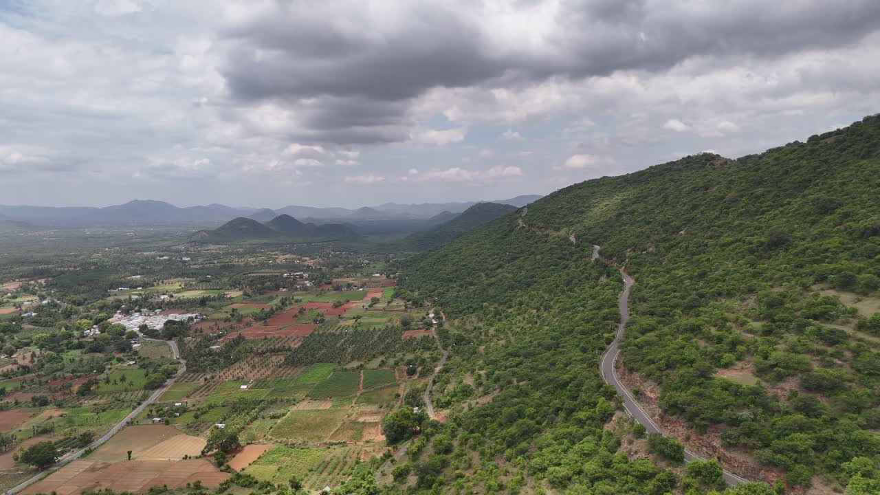 Stunning aerial view of a winding road with a series of hairpin turns cutting through a lush, green mountain valley scattered trees, leading up to densely forested hills in the background. Clouds