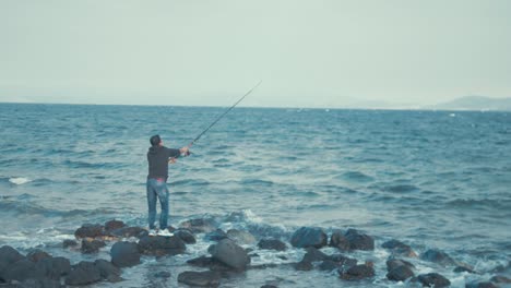 young man casting fishing rod standing on rocks at sea shore wide shot