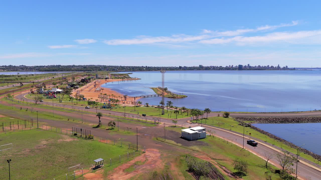 Balneario Costa Sur, Posadas with sandy beach, palm trees, and public park on the Paraná River, viewed by drone with city skyline beyond under a clear sky