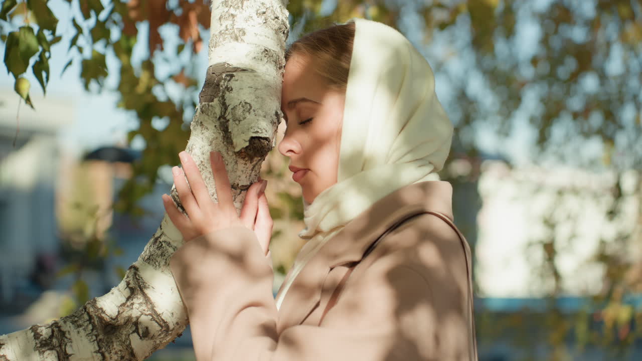 Donna bianca con foulard che tocca una betulla, passeggiata in un parco urbano in pausa, luce solare screziata sulle foglie, caldo cappotto color cammello, primo piano intimo, connessione tattile con la natura, atmosfera autunnale nostalgica, rallenty cinematografico.