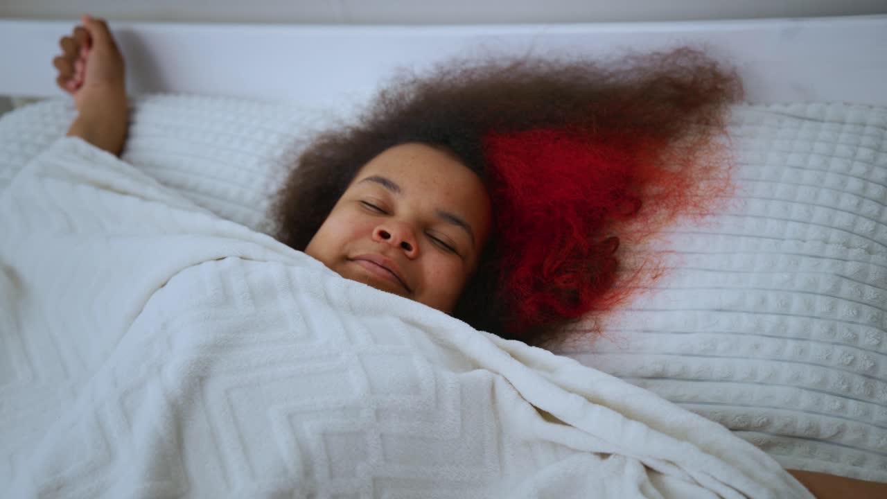 Cheerful young woman with curly red hair lazily stretching under soft white blanket, enjoying peaceful morning moments in cozy bedroom setting