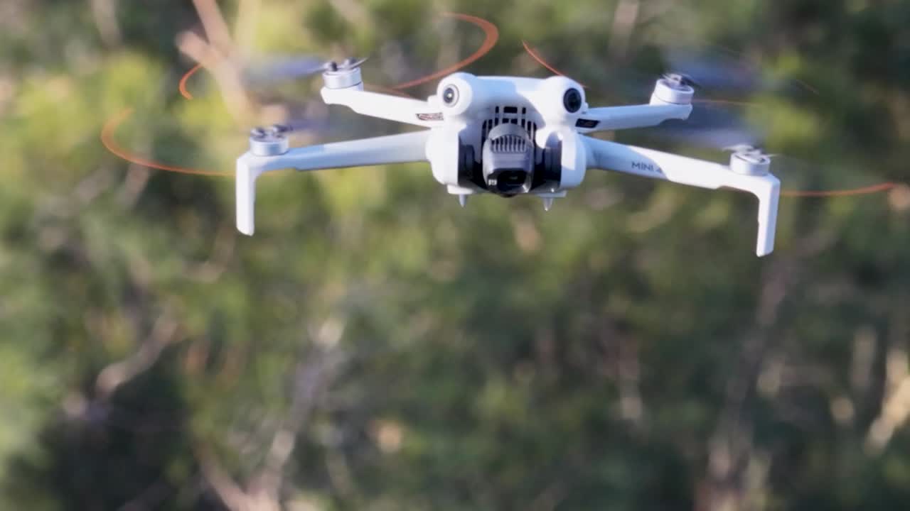 Un drone quadricottero bianco si libra stabilmente a mezz'aria con fogliame verde sfocato sullo sfondo.