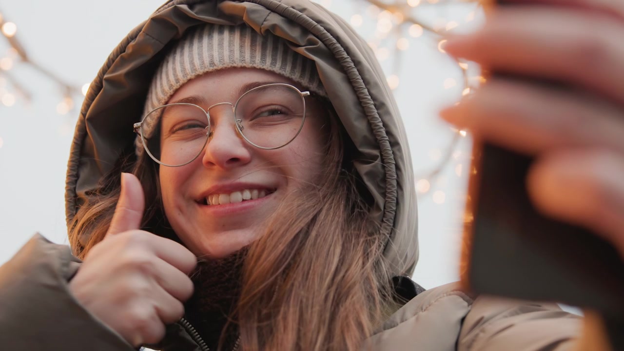Jonge witte vrouw maakt selfie buiten, draagt muts en jas met capuchon, ronde bril reflecteert kerstverlichting, speelse poses omvatten vredesteken, brede glimlach, zachte pruillip vanuit close-up hoeken.