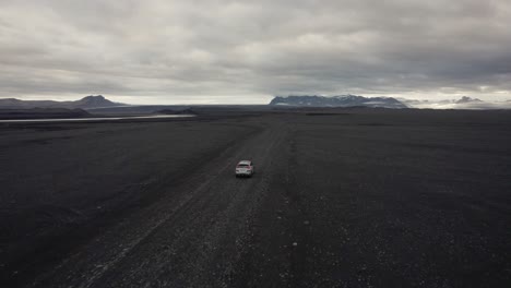 Drohnenaufnahme-Eines-Silbernen-Autos,-Das-Auf-Der-Straße-In-Einem-Lavafeld-In-Island-Fährt,-Mit-Kreisender-Kamera-Und-Bergen-Im-Hintergrund-4k