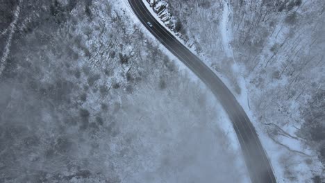 Flying-through-clouds-over-snow-covered-mountains-and-forests-in-a-mountain-valley-with-a-scenic-highway-below,-on-a-cloud-winter’s-day