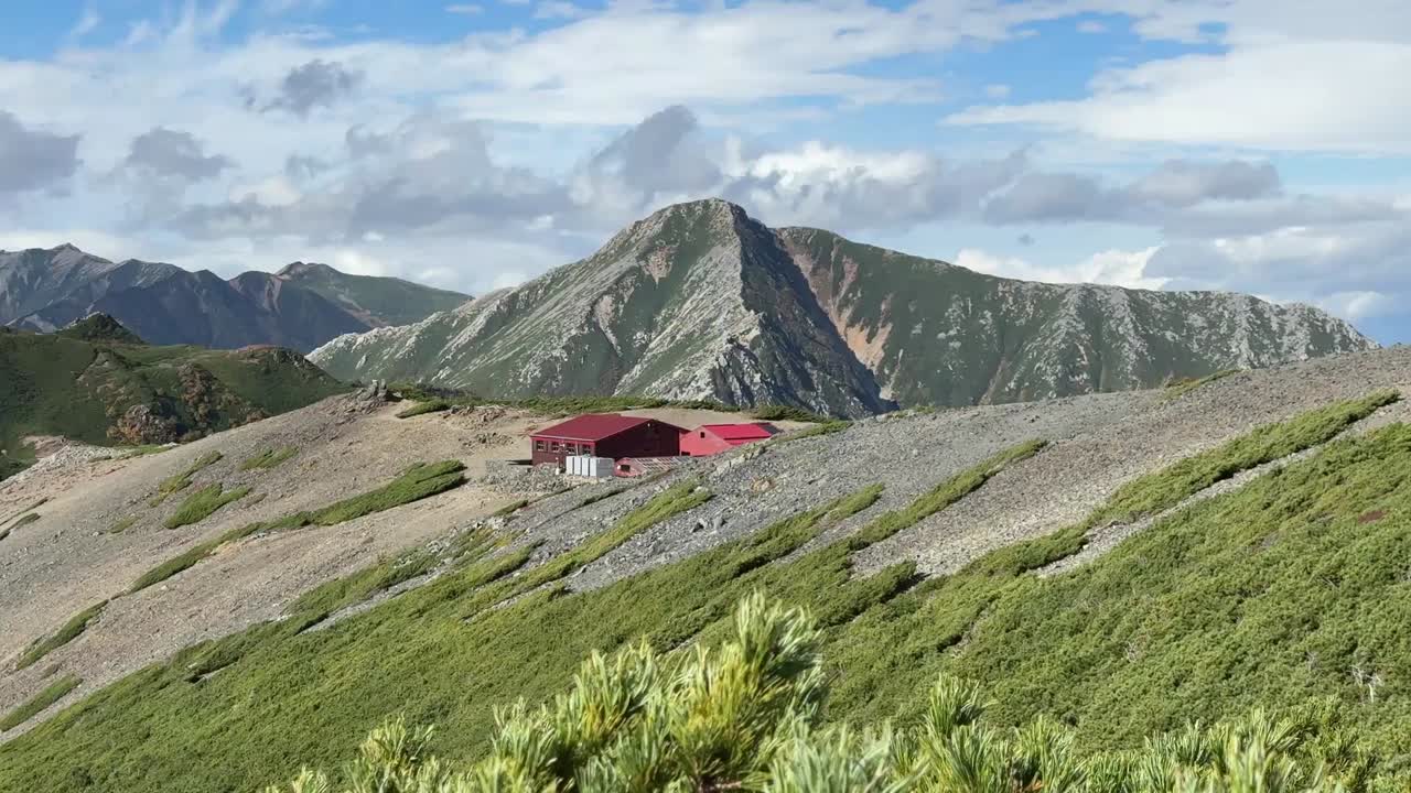Ampia inquadratura del rifugio Chogatake su un pendio alpino roccioso con le ripide montagne Hotaka che si ergono sullo sfondo sotto un cielo luminoso