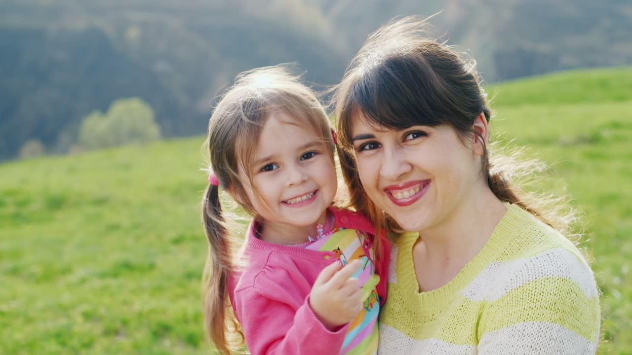 Una niña pequeña con dos coletas divertidas abraza a su madre y miran juntas a la cámara con alegría.