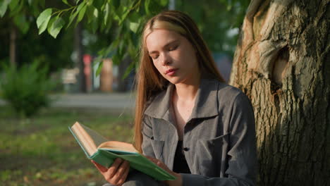 lady in grey clothing seated outdoors reading, looking thoughtful, the background features greenery and blurred building, with leaves above swaying gently in the wind