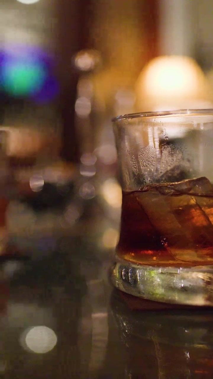 Bicchiere di whisky con ghiaccio su un bancone riflettente, illuminazione ambientale calda, messa a fuoco ridotta, movimento lento della telecamera.