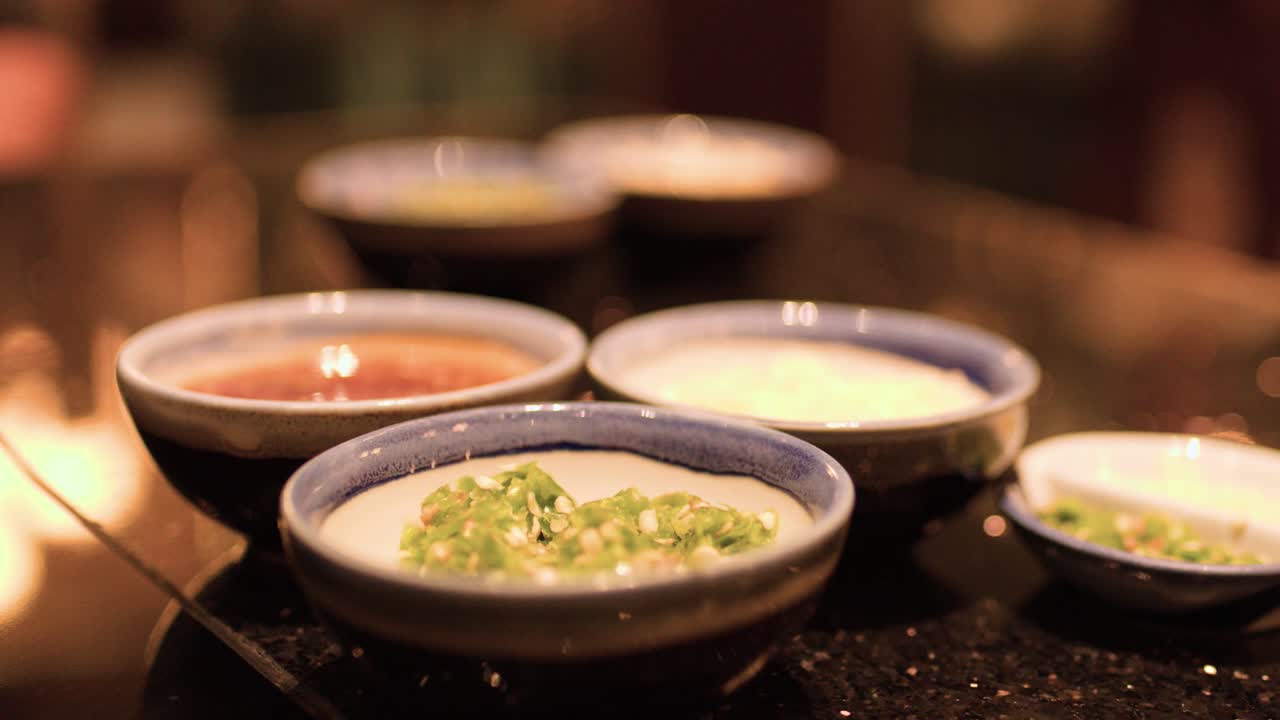 Three ceramic bowls with chili, garlic, and soy sauces on glossy restaurant table, warm lighting