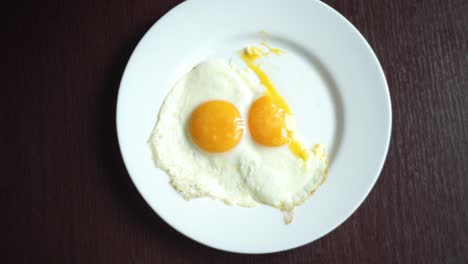 Cutting-fried-eggs-with-knife-and-fork-on-white-plate-at-wooden-table