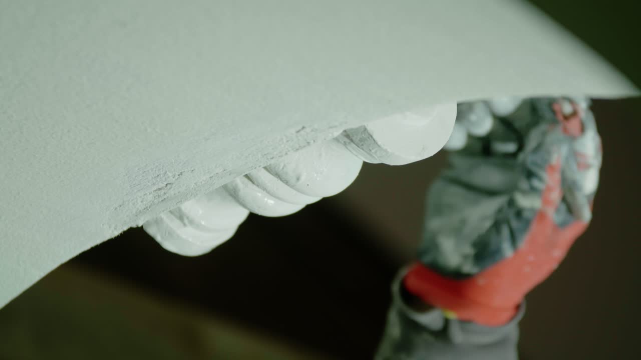 Extreme close-up, static shot of a worker's gloved hand using a small brush to carefully paint or touch up the intricate, white, egg-and-dart pattern on decorative architectural molding