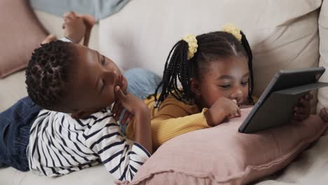 happy african american brother and sister using tablet relaxing on sofa at home, slow motion