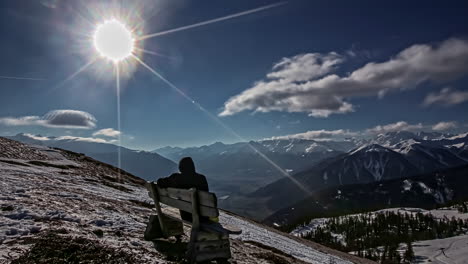 time lapse shot of person sitting on bench of snowy mountaintop and enjoying mountain panorama view during sunny day on blue sky in winter