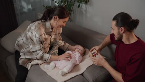 Happy-brunette-girl-in-a-plaid-shirt-together-with-her-brunette-husband-in-a-red-T-shirt-play-with-their-little-daughter-using-a-variety-of-toys-for-children-as-a-baby-in-a-pink-overalls-sitting-on-a-gray-sofa-in-a-modern-apartment