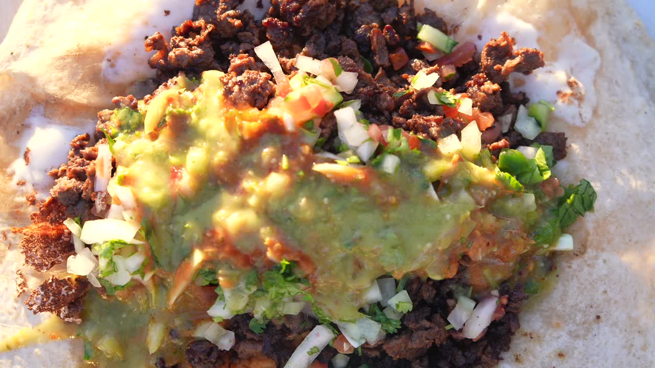Riprese ravvicinate di un burrito ripieno di carne, salsa messicana, cipolla e coriandolo presso una bancarella di street food di taco. Il lento movimento della telecamera evidenzia la consistenza, la freschezza e la presentazione autentica del cibo