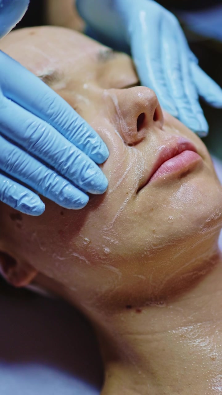 Una donna riceve un trattamento viso in una spa di giorno, indossando una fascia per capelli, mentre mani guantate le applicano prodotti sulla pelle.