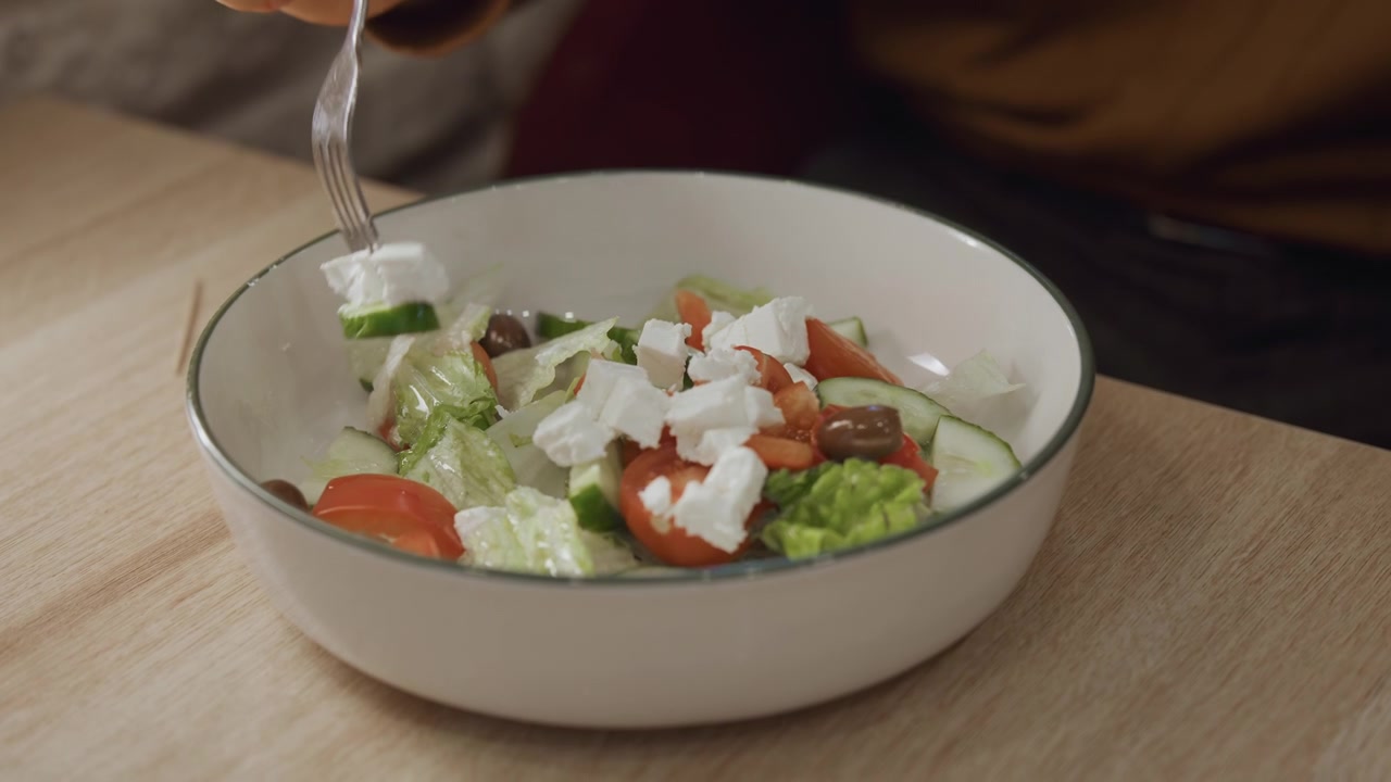 Plano detalhe da mão de uma pessoa usando um garfo para pegar ingredientes frescos em uma tigela contendo folhas de salada, tomates, pepinos, azeitonas e queijo esfarelado. A cena apresenta iluminação suave sobre uma mesa de madeira.