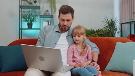 Child-kid-daughter-having-fun-with-father-or-nanny-enjoying-using-laptop-pc-watch-cartoons-at-home