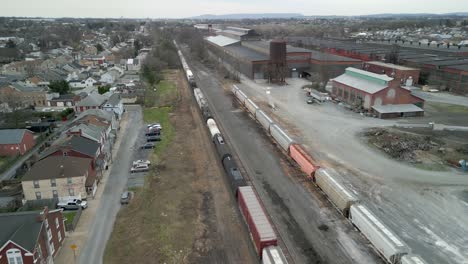 Aerial-drone-view-of-train-traveling-down-a-railway-with-an-industrial-plant-on-one-side-and-housing-on-the-other