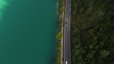 a car drives along a road beside a turquoise lake and lush green forest, shot from above