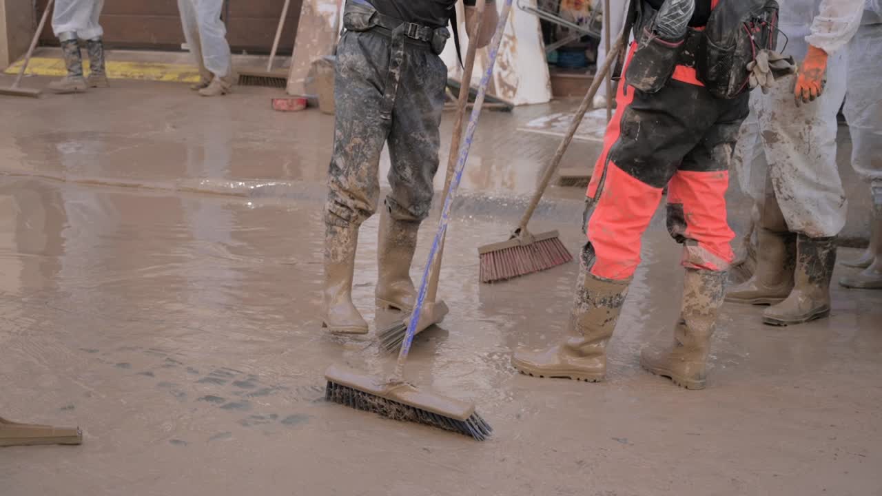 Workers wearing boots and protective gear clean up muddy water with large brushes after a flood in Valencia