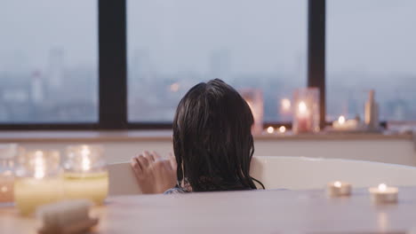 rear view of a woman taking a bath in a bathtub while looking out the window, then looks at the camera and smiling