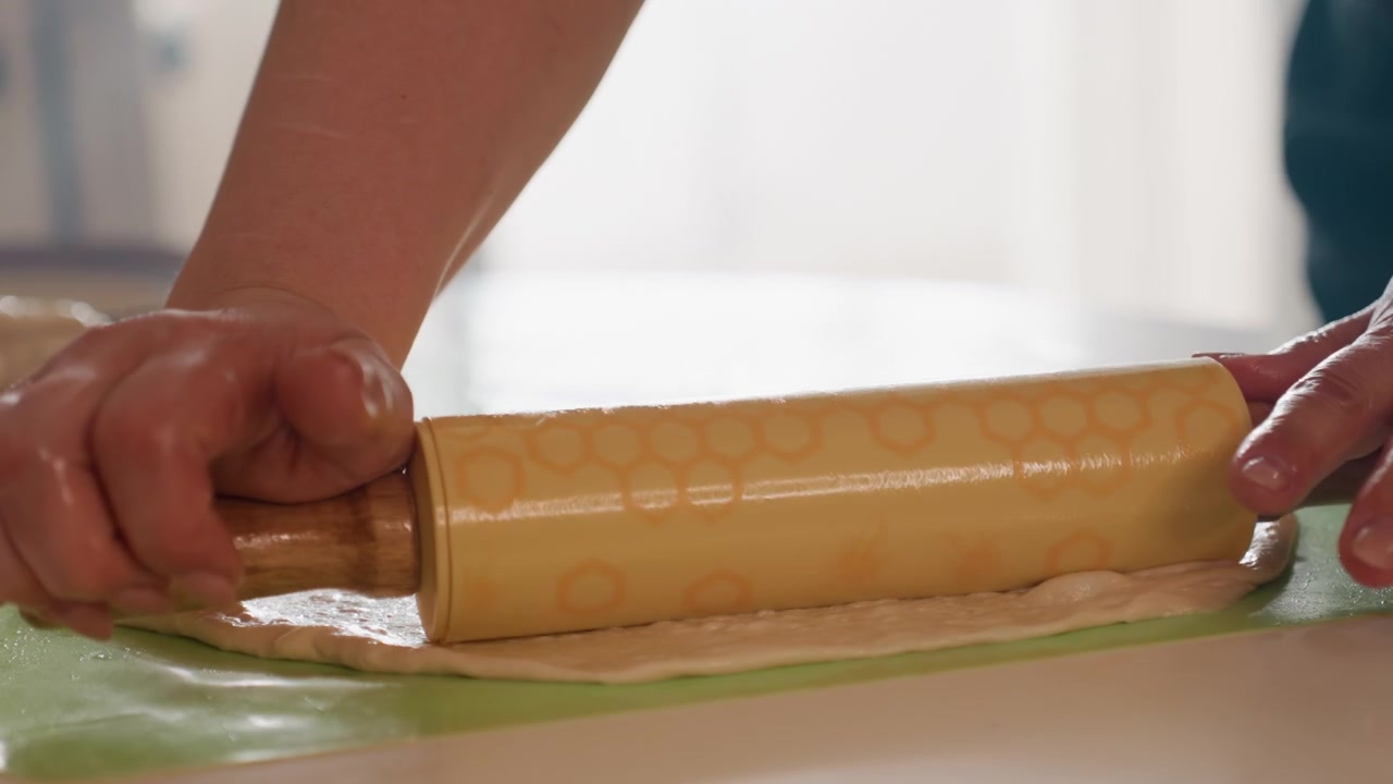 Nahe Aufnahme aus der Froschperspektive von Händen, die Teig auf einer grünen Silikonmatte bearbeiten. Ein hölzernes Nudelholz bewegt sich über die weiche Oberfläche, während der Teig sich dehnt, flach wird und sich zum Backen sammelt.