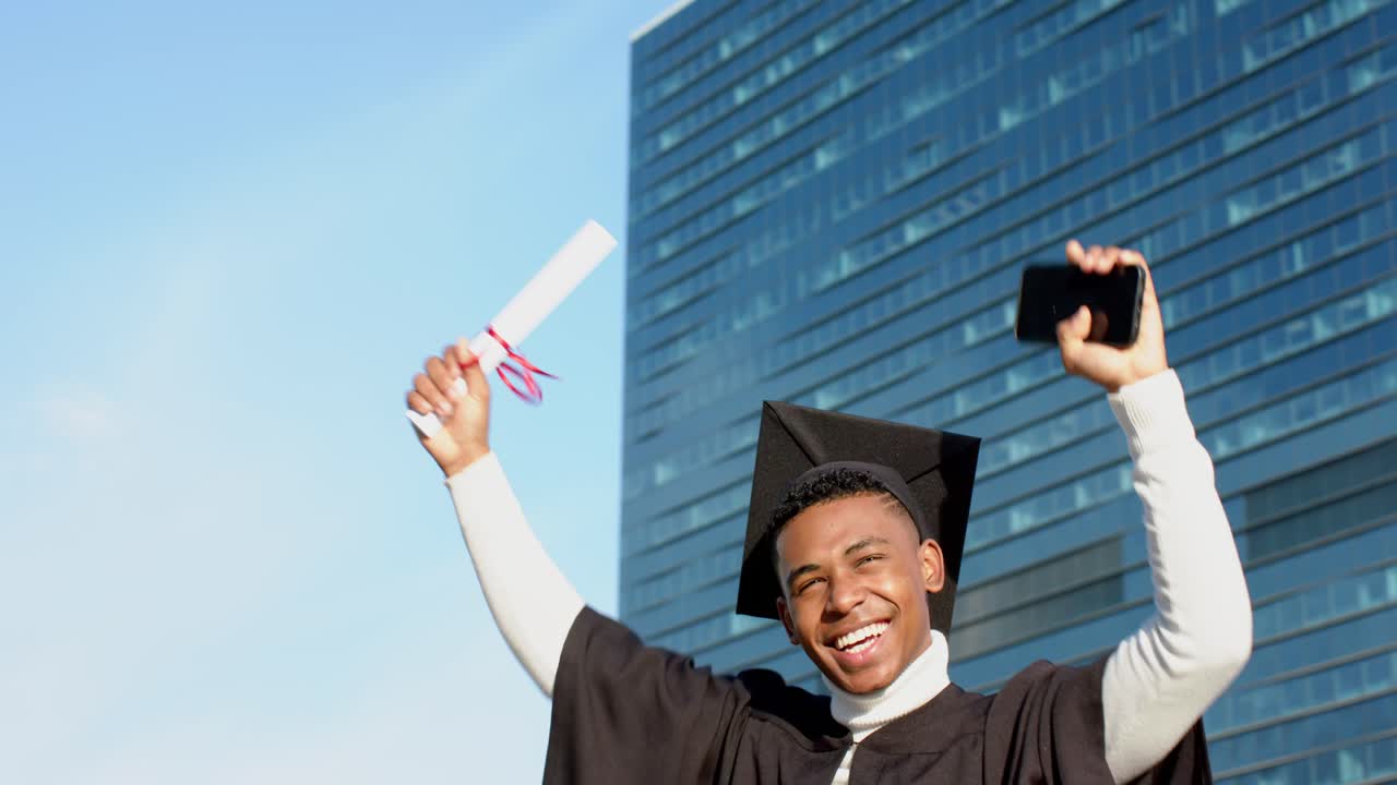 African American man holding diploma and smartphone at plaza celebrating graduation posting success