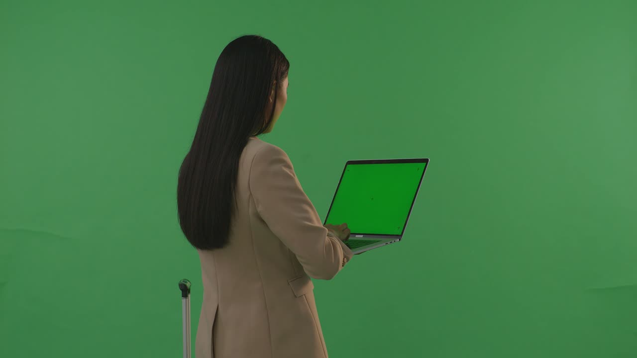 vista trasera mujer de negocios asiática viajera con equipaje usar portátil de pantalla verde mientras está de pie en el estudio de fondo de pantalla verde, durante la hora de la puesta del sol