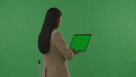 back view asian businesswoman traveler with luggage use green screen laptop while standing in the green screen background studio, during sunset time