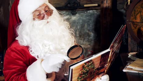 santa claus is reading at home. real santa in red and white costume wearing glasses looking into a book through magnifying glass at christmas time