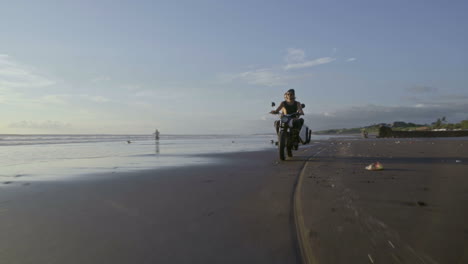 couple riding a motorcycle carrying a surfboard on the beach