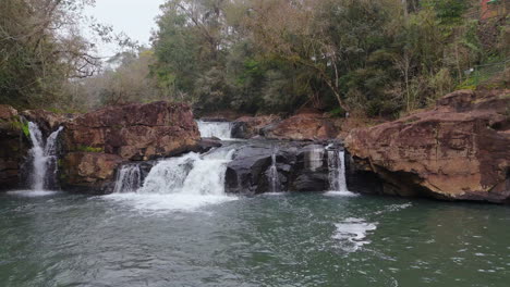 stunning drone footage captures waterfalls flowing into a river amidst lush green vegetation and rocky terrain, creating a picturesque natural landscape, low altitude drone pan
