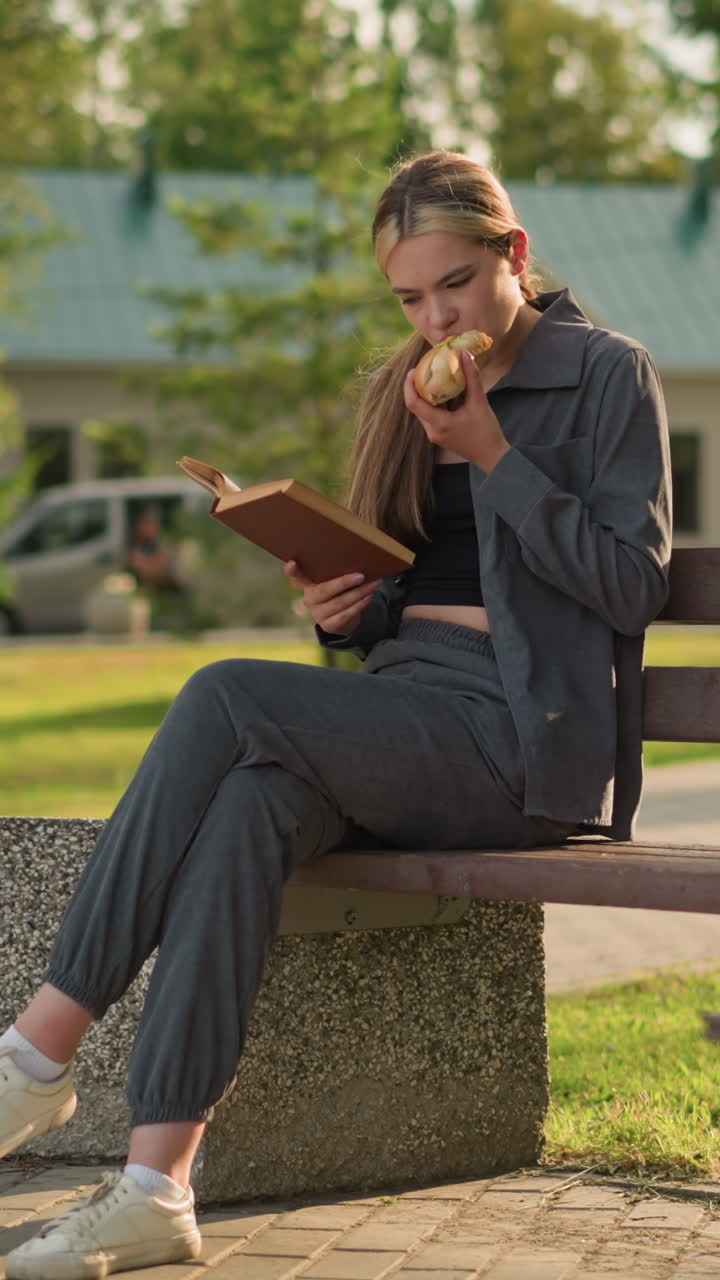 Lady seated on park bench eating burger while reading outdoors, with two people walking in the background and a relaxed park setting, with trees, benches, and a sunny atmosphere