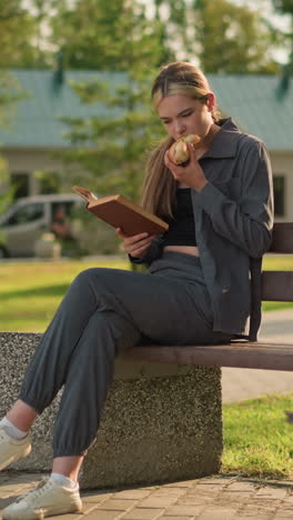 dama sentada en un banco del parque comiendo una hamburguesa mientras lee al aire libre, con dos personas caminando en el fondo y un entorno de parque relajado, con árboles, bancos y una atmósfera soleada