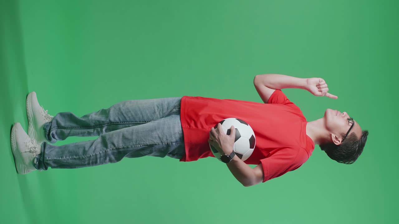 눈이 닫힌 몸으로 가득 찬 아시아 남자가 공을 들고 지표손가락을 올리고 머리를 흔들며 녹색 화면 배경에서 축구를 응원합니다.