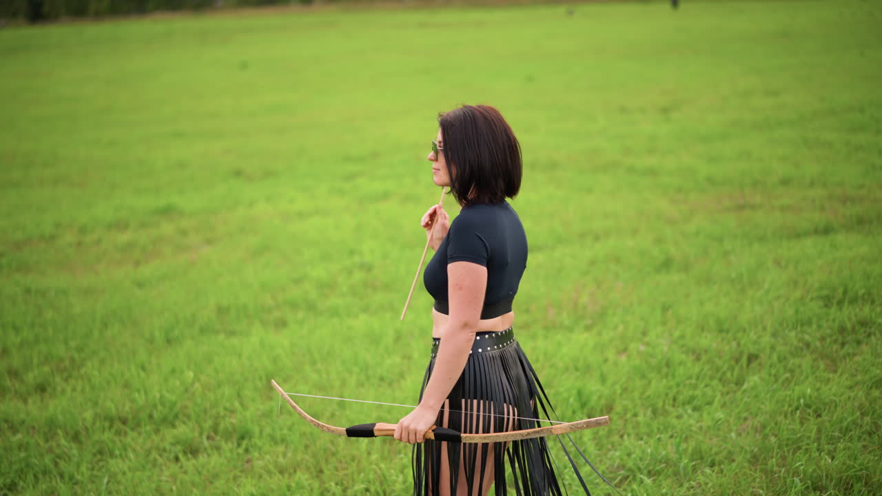 introverte boogschuttervrouw met pijl in de hand, klaar om te spannen in een weide, casual crop top en rok met franjes en boog op de heup, intieme profielfoto's leggen de vaste greep vast, zonnebril, kalm groen gras