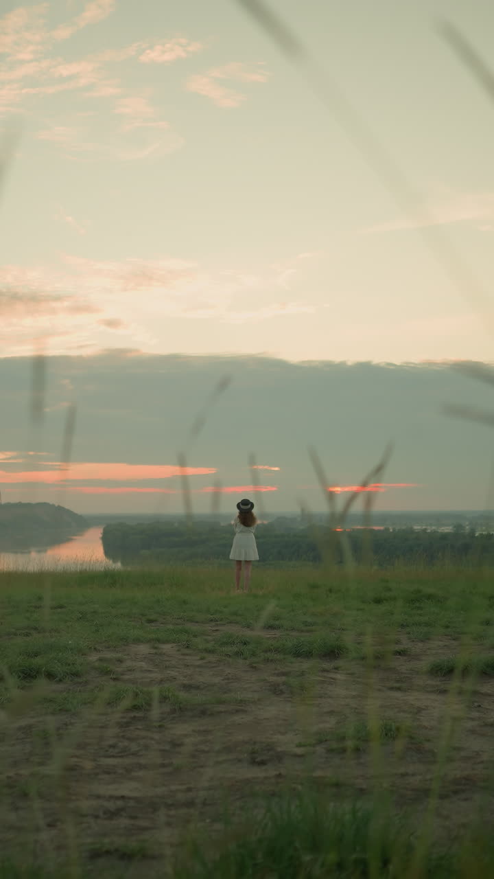 una vista distante de una mujer con un vestido blanco y un sombrero, de pie pensativa en un campo cubierto de hierba por un lago tranquilo al atardecer. ella mira hacia el horizonte, abrazando el momento sereno y reflexivo