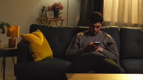 young man spending evening at home sitting on sofa with mobile phone scrolling through internet or social media 7