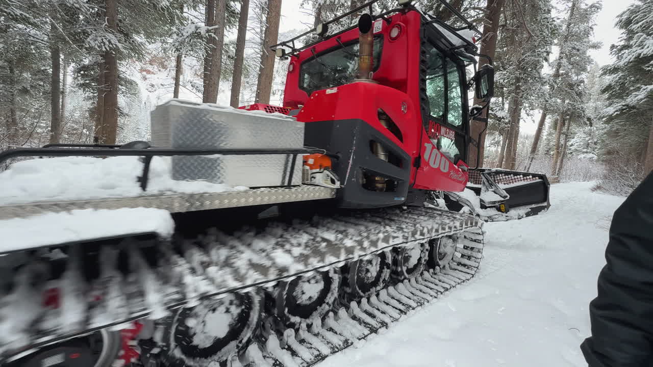 Un uomo con una giacca invernale scura cammina davanti a un gatto delle nevi rosso su una strada innevata nella foresta, mostrando attrezzature pesanti che mantengono l'accesso alla montagna nel cuore dell'inverno