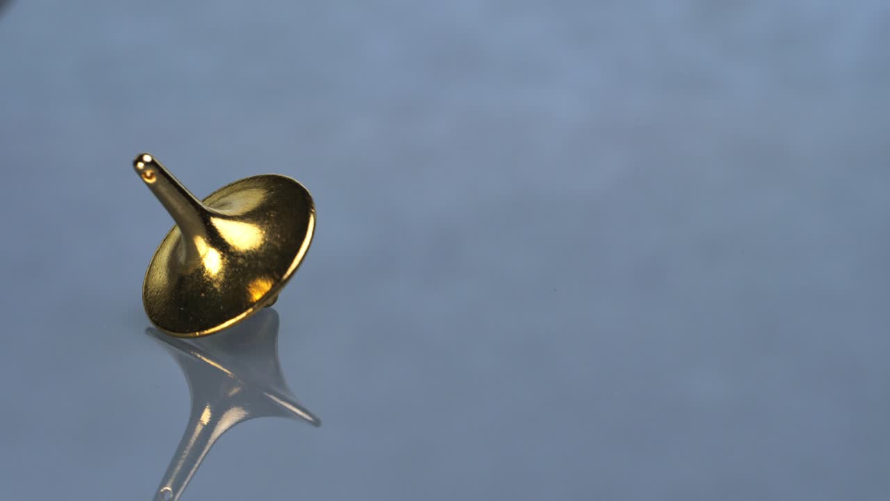 A golden, polished, spinning top wobbles on a reflective surface with a mirrored reflection and copy space