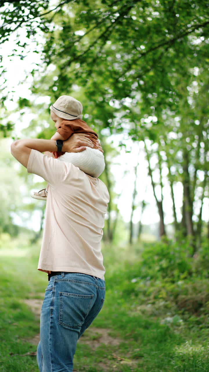 Caucasian man hops holding his toddler son on the shoulders. Father entertains his child outdoors on the walk in summer. Vertical video.