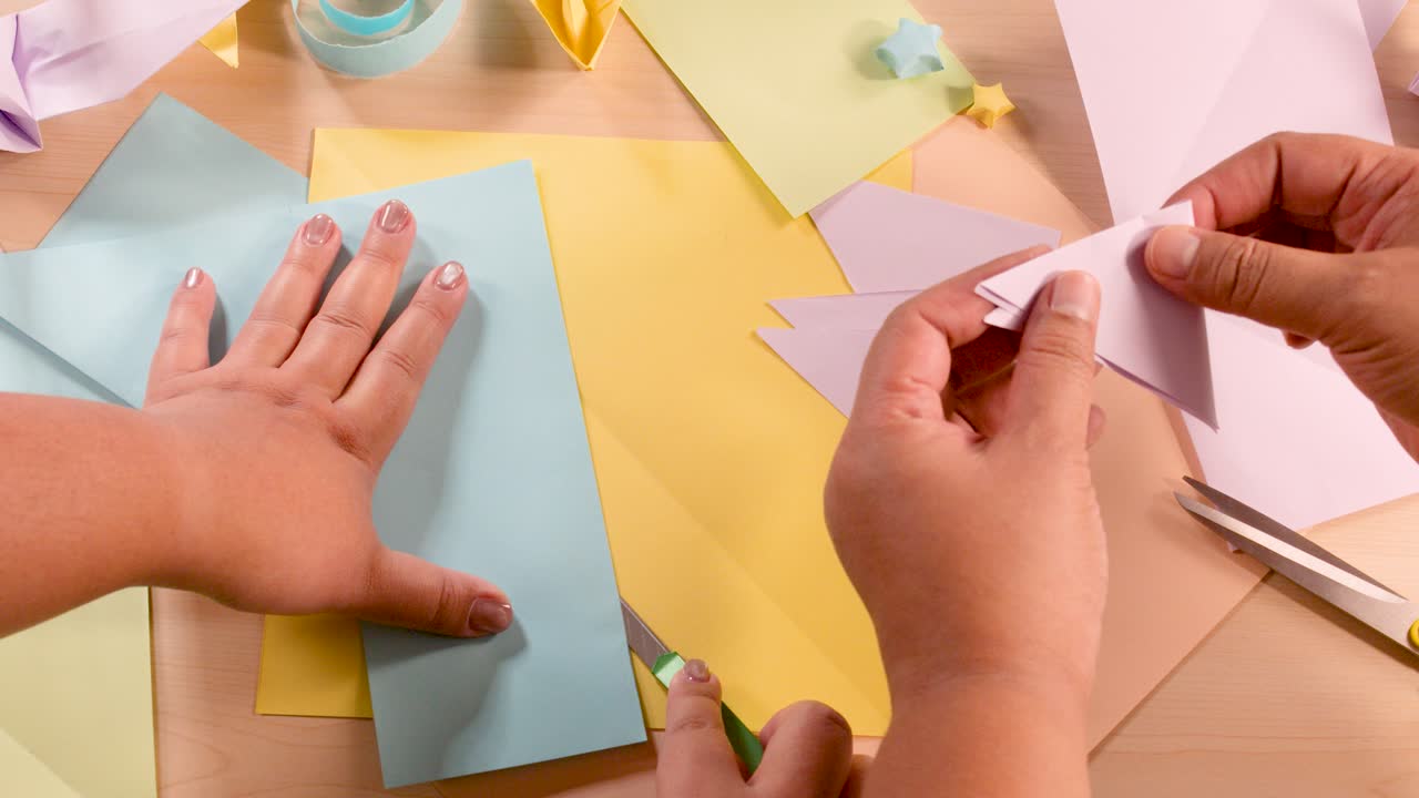 Person folds pastel origami paper on organized craft table, using scissors, under soft, even lighting
