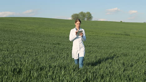 Caucasian-researcher-woman-in-white-coat-walking-in-green-field-and-a-using-tablet